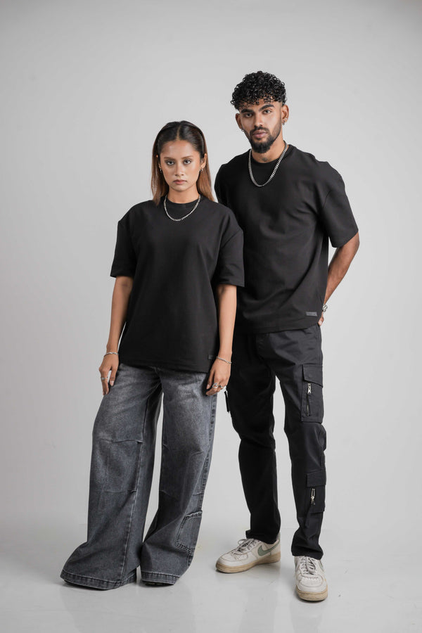 Two people wearing black oversize  t-shirts and dark jeans standing against a gray background