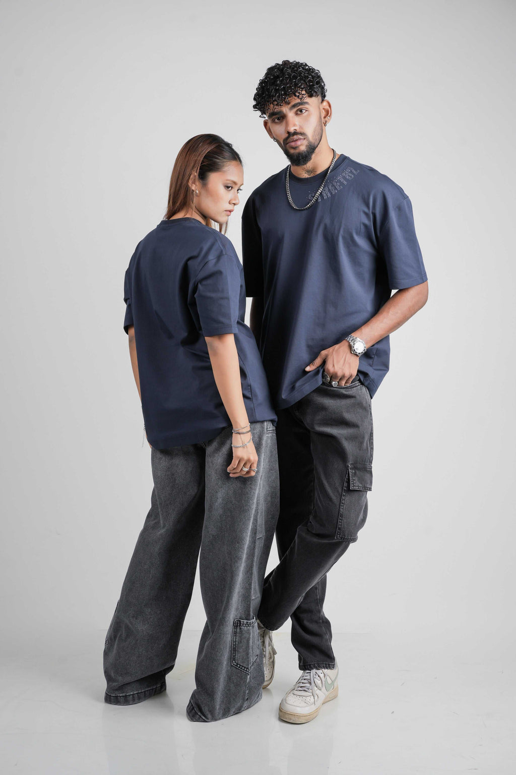 Two people wearing matching navy blue oversize t shirts and dark pants on a plain background